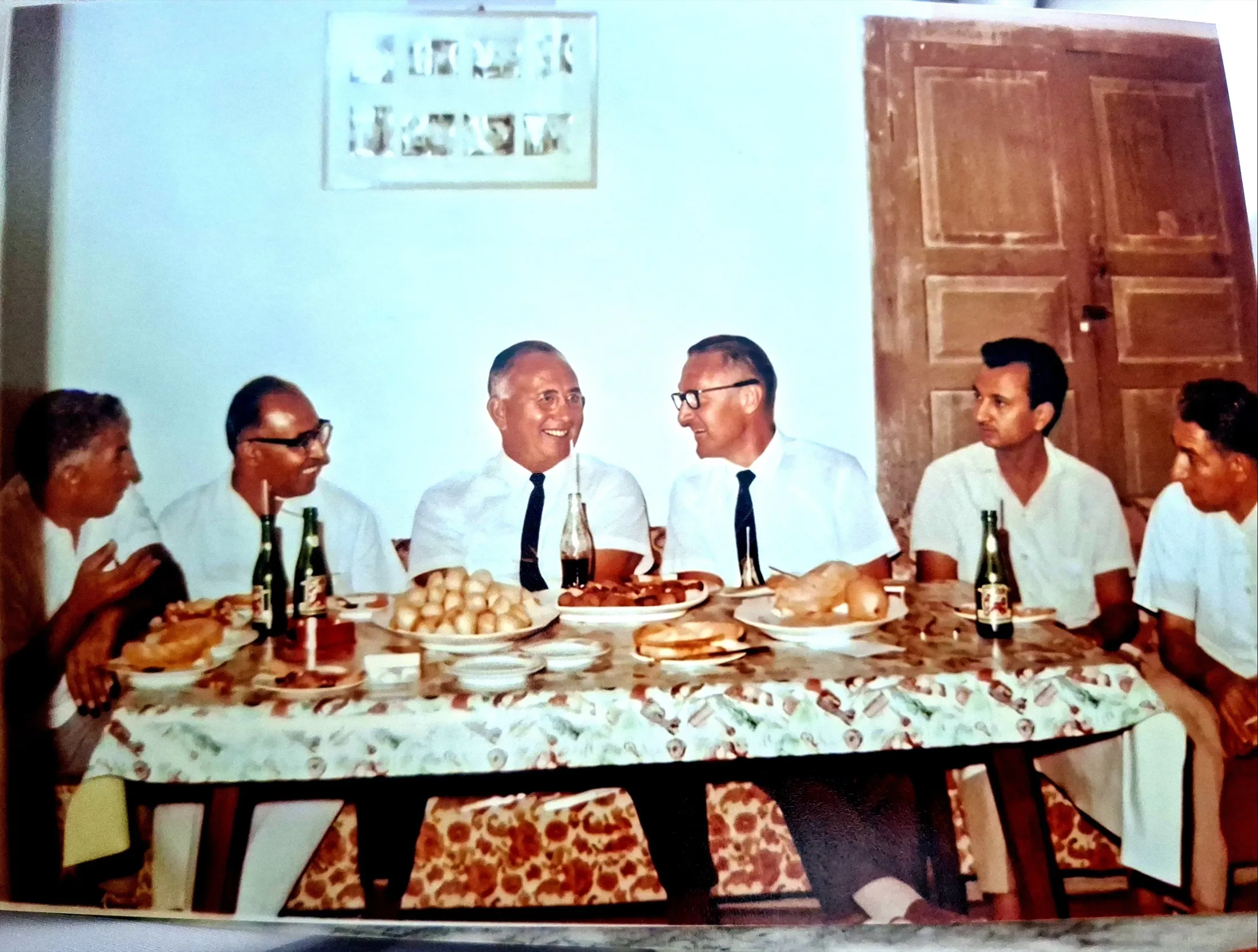 Men seated at a long table laid with food and bottled drinks; two foreign guests laugh at the centre.