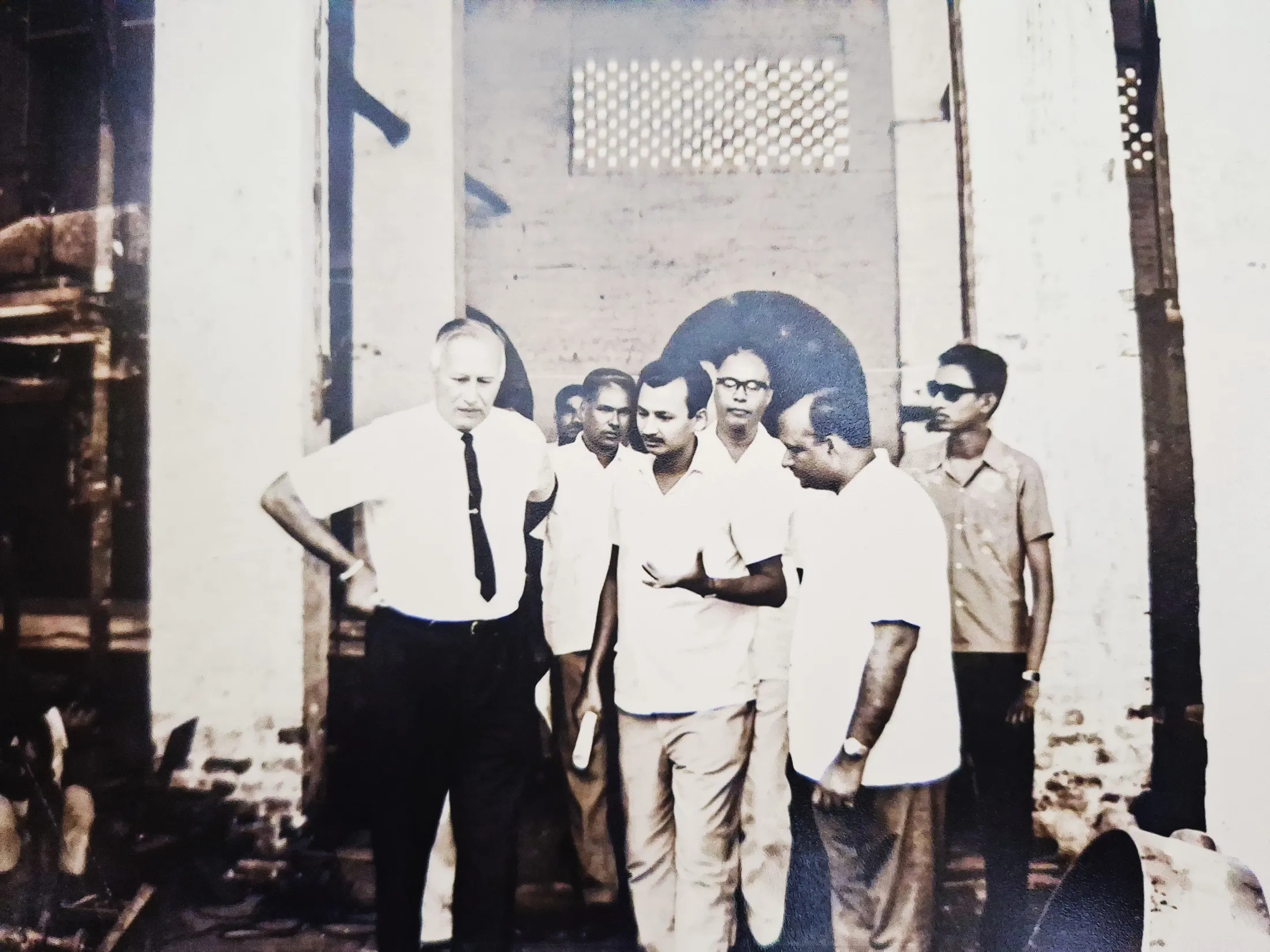 An older foreign engineer in shirt and tie talks with the management team inside the plant.