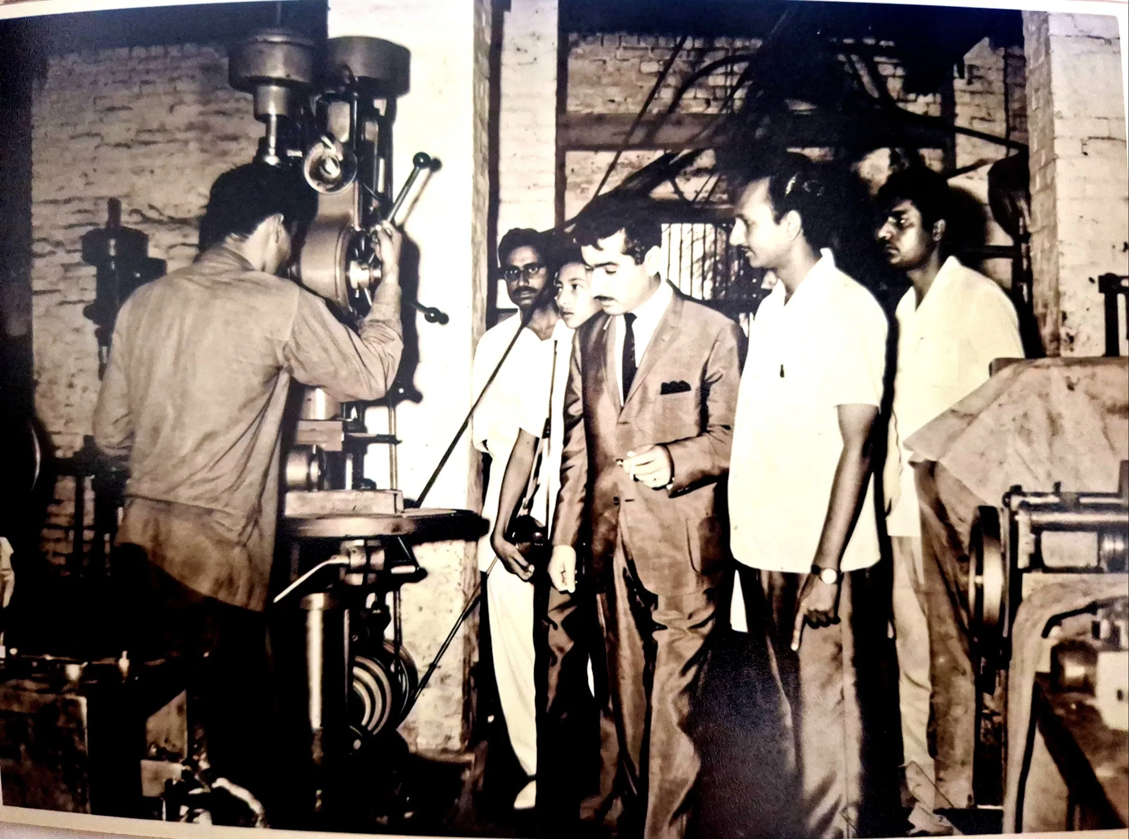 A fitter works a belt-driven drill press in the mill's machine shop while visitors watch.