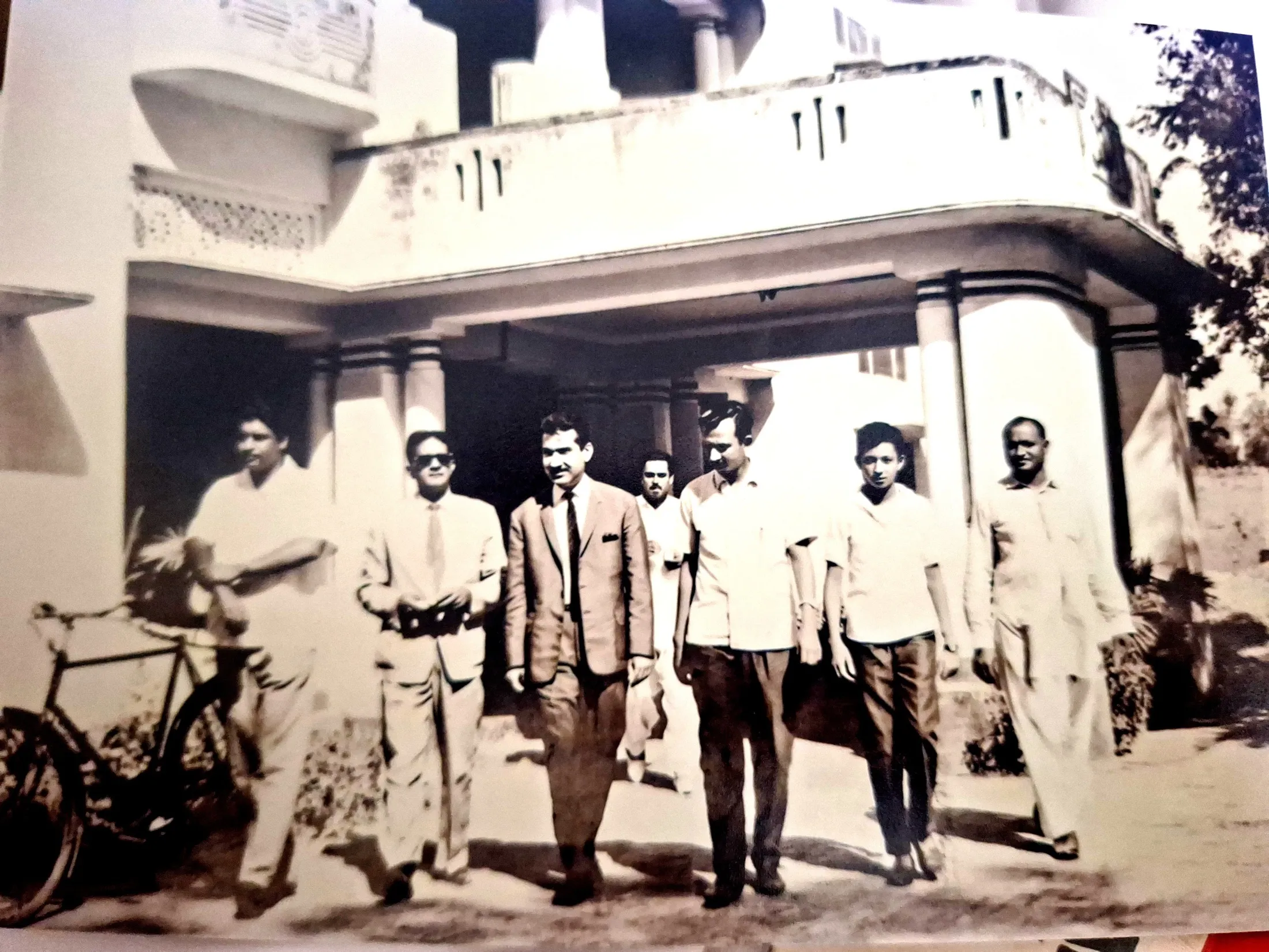 Men gathered outside a two-storey house with a columned veranda; a bicycle leans by the steps.